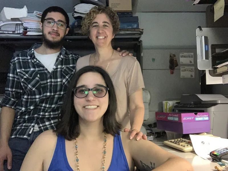 Three people smiling at the camera. A woman is seated in front wearing glasses. Two other people stand behind her in a cluttered office space.