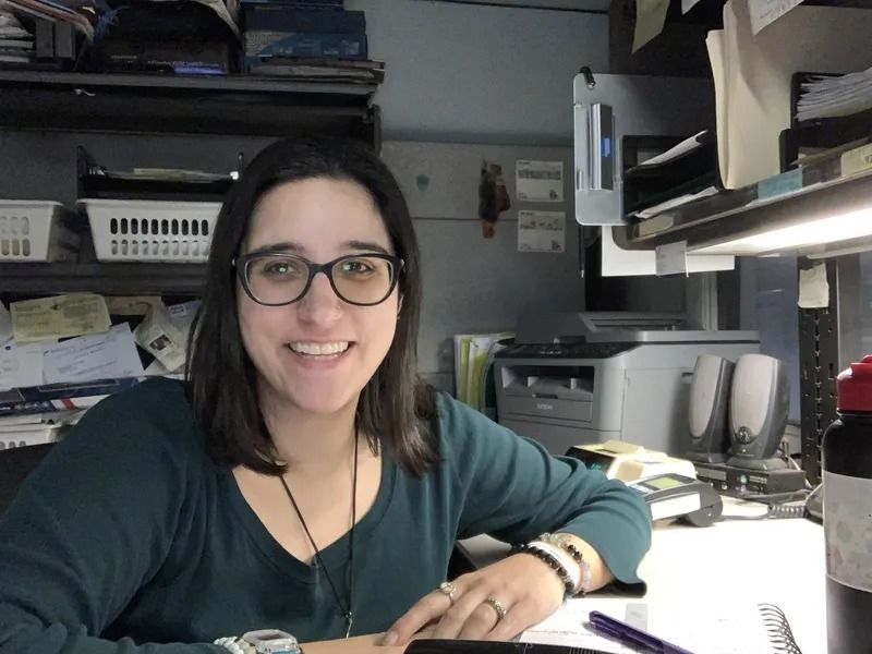 Woman with glasses smiles at the camera while seated at a cluttered office desk. She wears a green shirt.