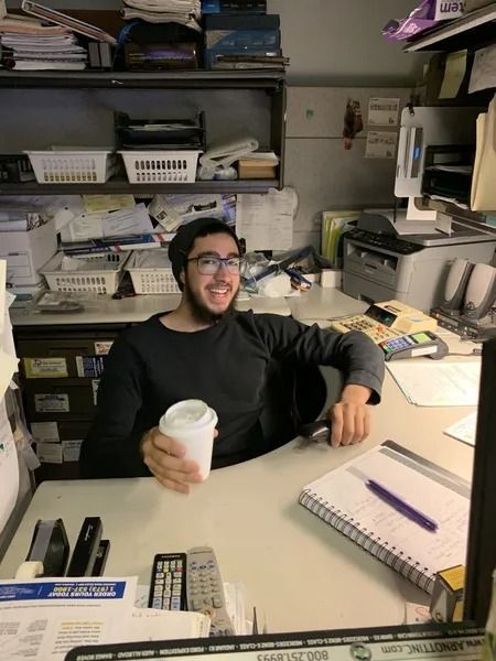 Man with a beard and glasses smiles while holding a coffee cup at a cluttered office desk.