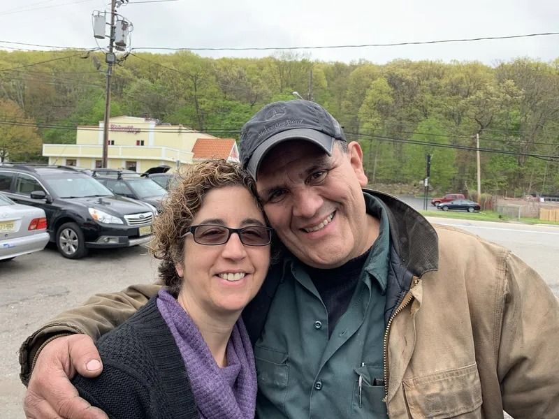 Two people smiling and embracing outdoors: a woman with curly hair and glasses, and a man with a baseball cap. They are in front of a parking lot with cars.