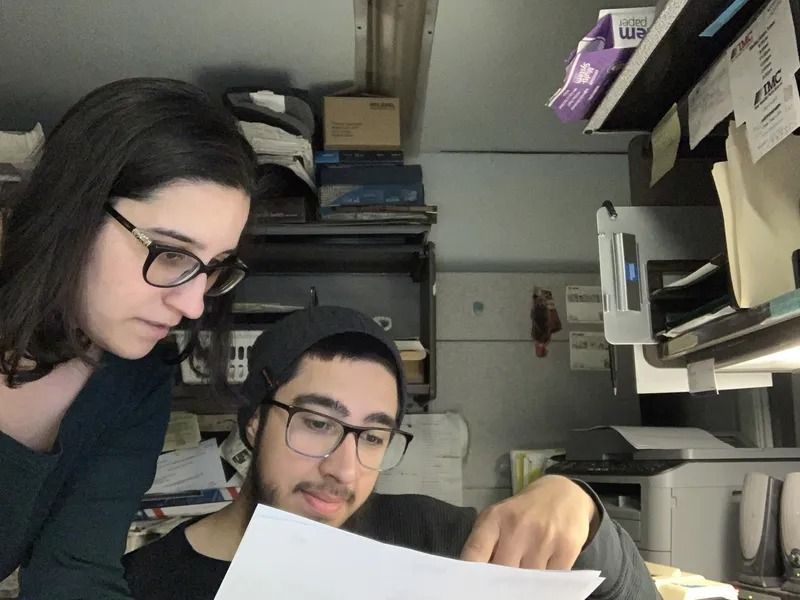 Two people, one woman and one man, looking at a paper together. The woman leans over the man's shoulder, both wearing glasses and focused. They are in an office environment.