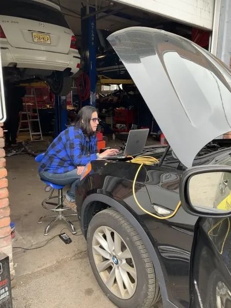 A person in a blue plaid shirt uses a laptop connected to a car's engine in a garage. The car's hood is open.
