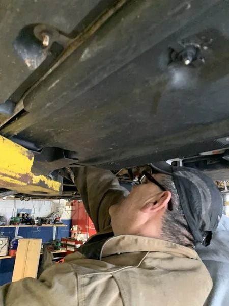 A mechanic in coveralls examines the undercarriage of a car raised on a lift. The setting is a garage.