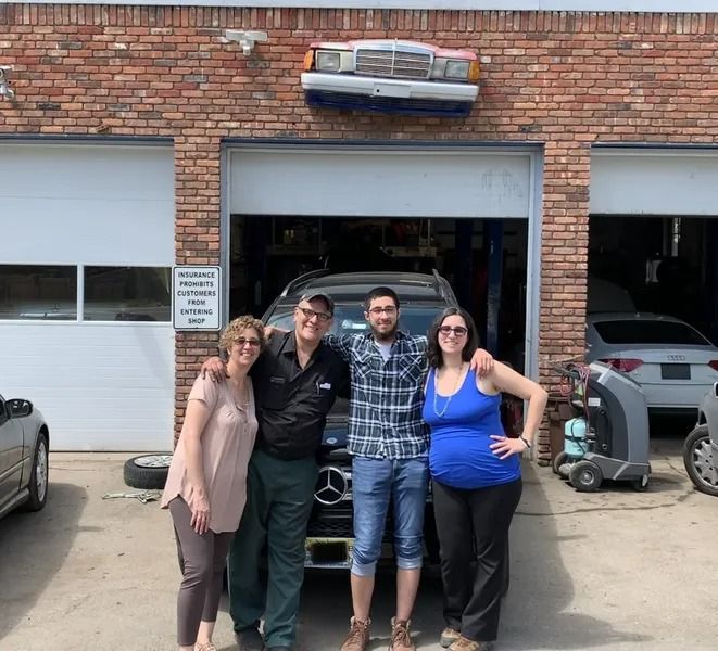 Four people pose in front of a garage, a Mercedes-Benz visible inside. A car hood decorates the top of the building.