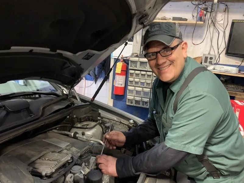 Mechanic in a green uniform and cap smiles while working on a car engine in a garage.