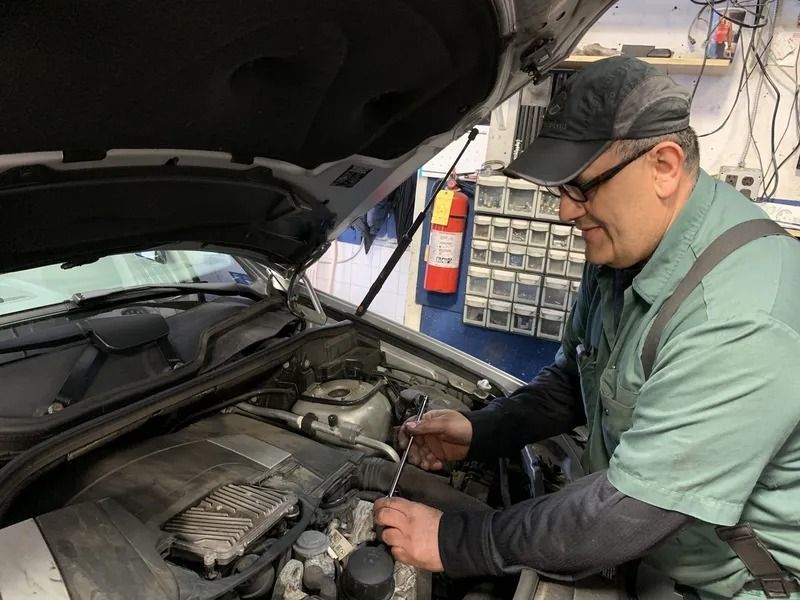 A mechanic in a green uniform works under the open hood of a car in a garage, examining the engine.