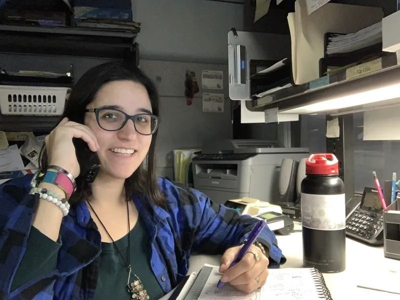 Woman in glasses smiles while on the phone and writing in a notebook at her desk, which has a water bottle and printer.
