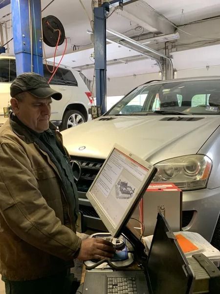 Mechanic in a garage, using a computer diagnostic tool on a silver car. He's wearing a brown jacket and hat. Another car is in the background.