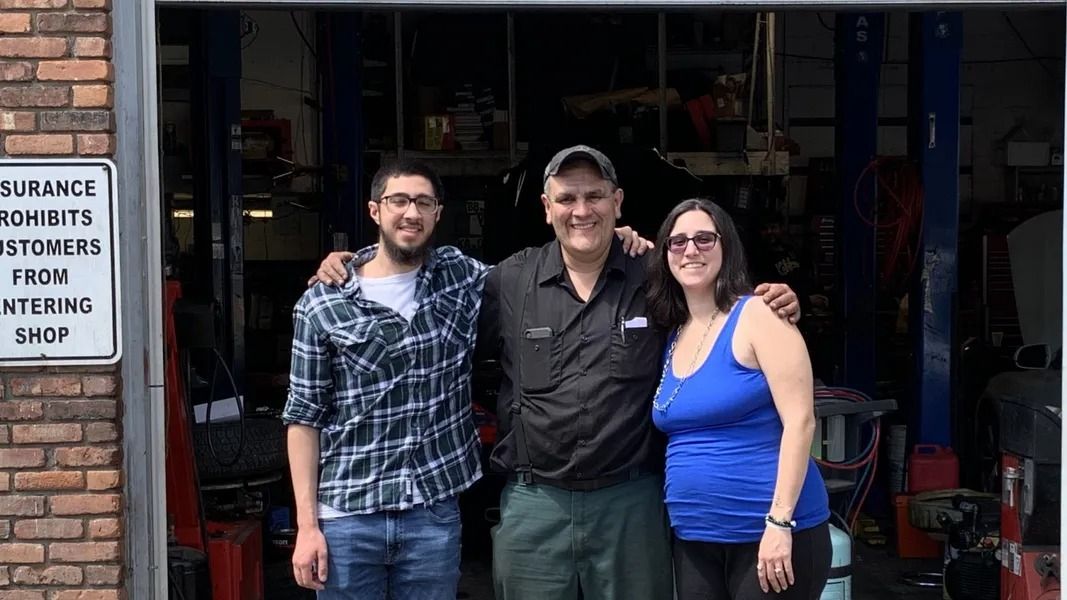 Three people stand in front of an auto shop, smiling at the camera. One is wearing a plaid shirt, one a black polo, and one a blue tank top.