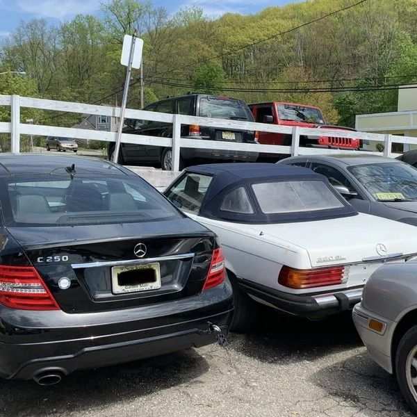 Cars parked in a lot, including a black Mercedes-Benz, a white convertible, and a red Jeep, with a white fence and trees in the background.