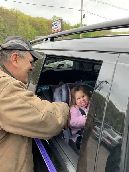 A man smiles at a young girl in a car seat; she sticks her tongue out, parked near a building with a sign.