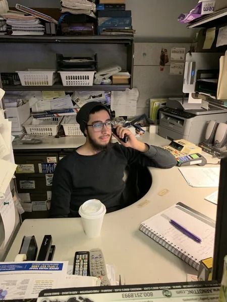 Man with glasses on phone at a messy desk; cup of coffee on the counter. Dark gray sweater, neutral background.