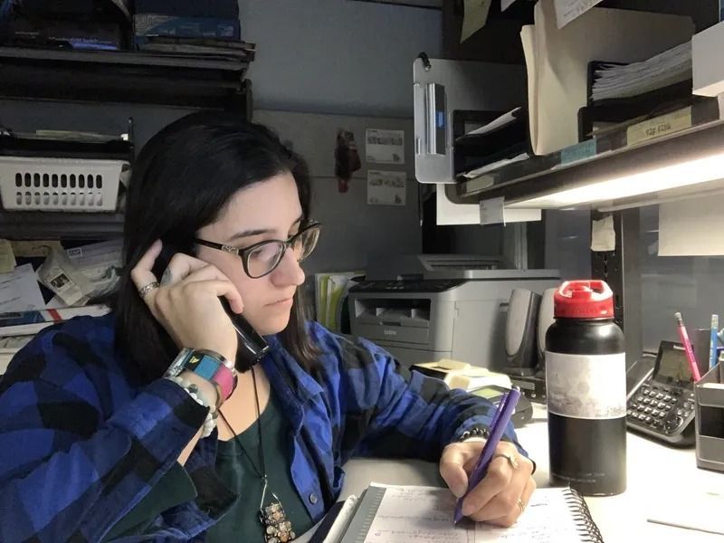 A woman with glasses in a blue plaid shirt speaks on the phone, taking notes in a notebook at her desk, illuminated by a fluorescent light.