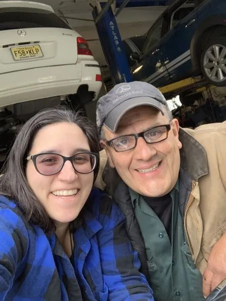 Two smiling people, a woman and a man, in an auto shop. The woman wears a blue plaid shirt and glasses, the man a hat, glasses, and a tan jacket.
