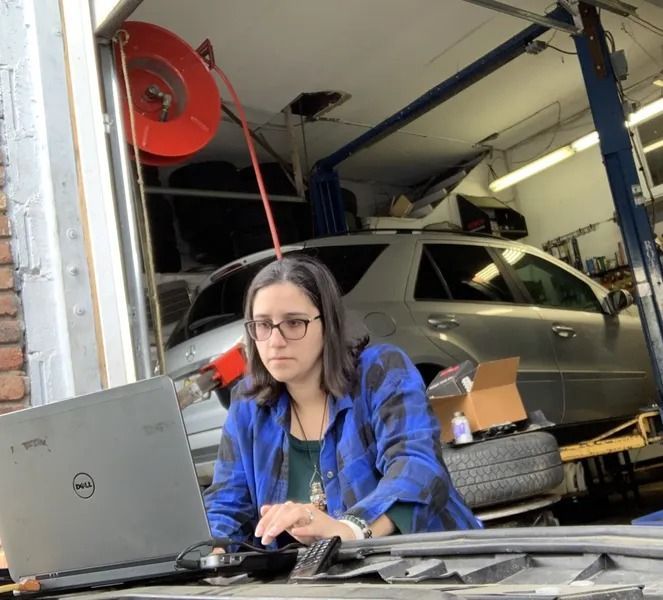 A woman in a blue plaid shirt and glasses works on a laptop in a garage, with a car in the background.