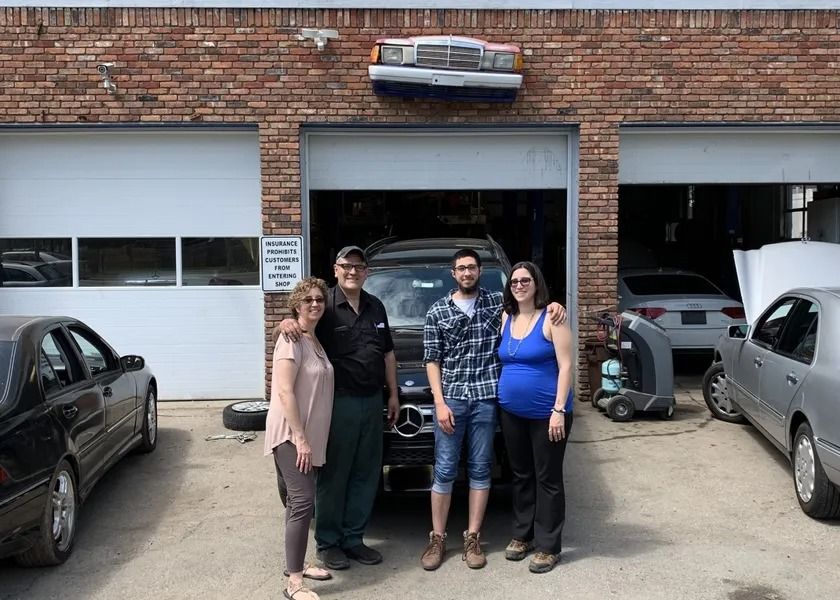 A group of five people pose in front of a car garage, with a classic Mercedes-Benz car. They're smiling. The garage has a half car facade.