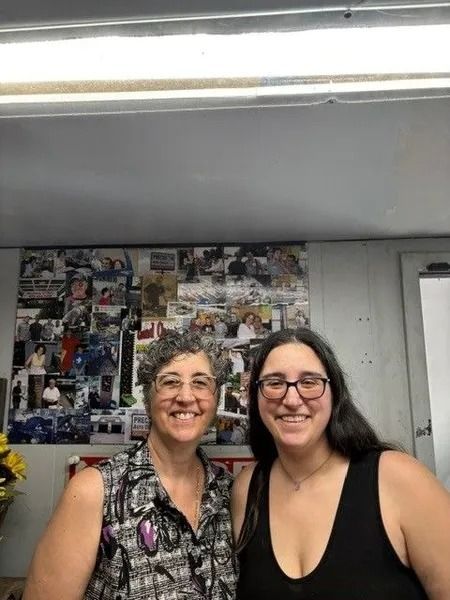 Two smiling women, one older with short hair and glasses in a patterned top, and one younger with long dark hair in a black tank top. They stand in front of a photo collage.