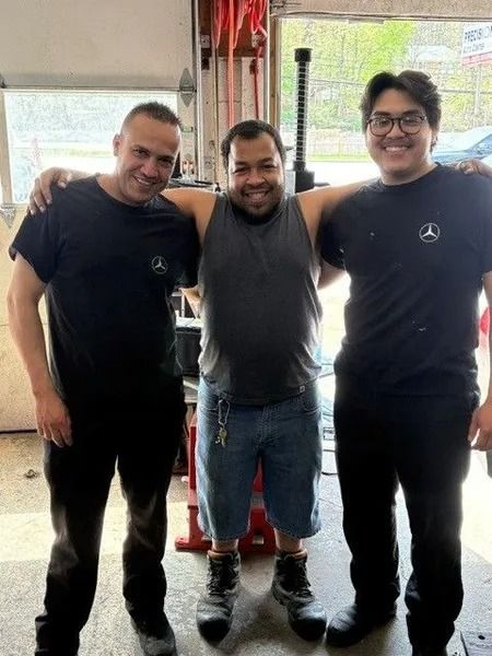 Three men in a garage, posing with arms around each other. They are smiling, wearing work clothes, and appear to be mechanics.