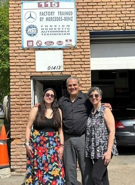 Three people pose in front of a brick building with a Mercedes-Benz auto repair sign. The setting is outdoors, on a sunny day.