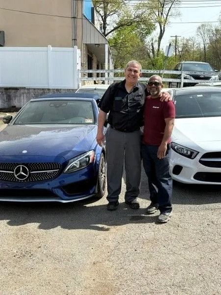 Two men stand next to two Mercedes-Benz cars. One car is blue and the other is white. They are smiling outside.