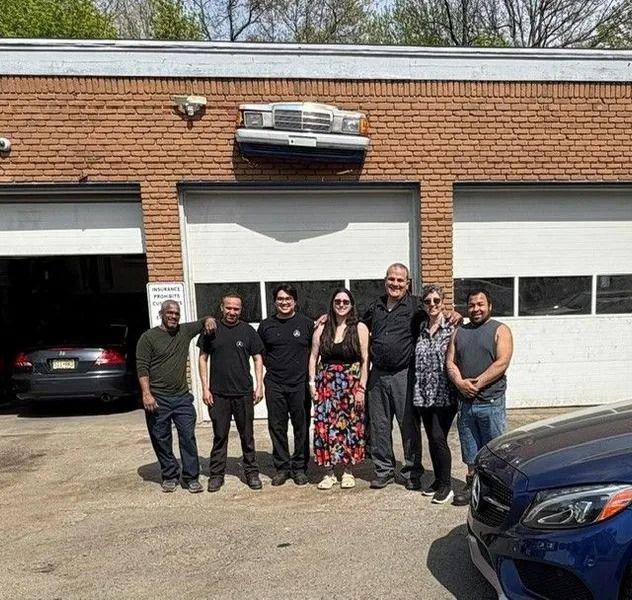 Group of people standing outside a brick auto repair shop with garage doors. People are smiling; cars are visible.