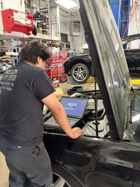 A mechanic working on a black car with an open hood, using a laptop. They are in a garage setting with other vehicles visible.
