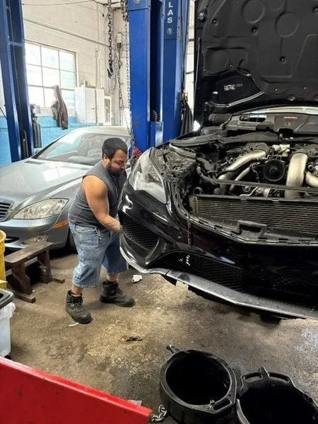 A mechanic in a garage works on a black car with its hood open. Another car is in the background.