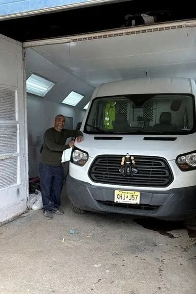 Man leaning on a white van inside a garage. The man is smiling and the van has a New Jersey license plate.