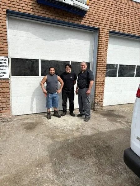 Three men stand in front of garage doors. They appear to be posing. The setting is a brick building, possibly an auto repair shop.