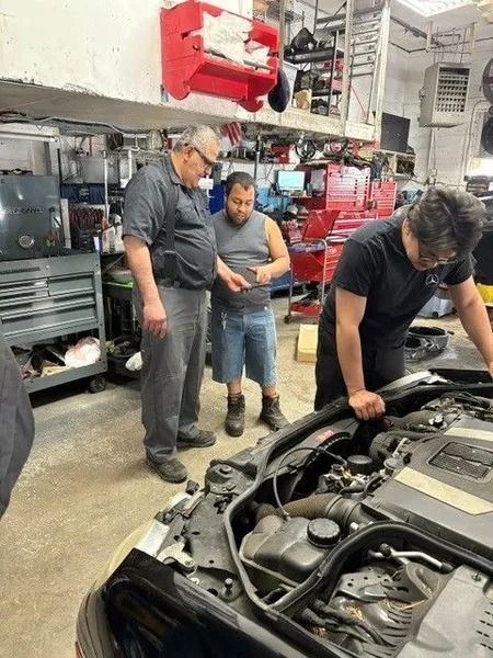 Three mechanics in a garage looking at a car engine. Two men are examining a phone, while another works on the car.