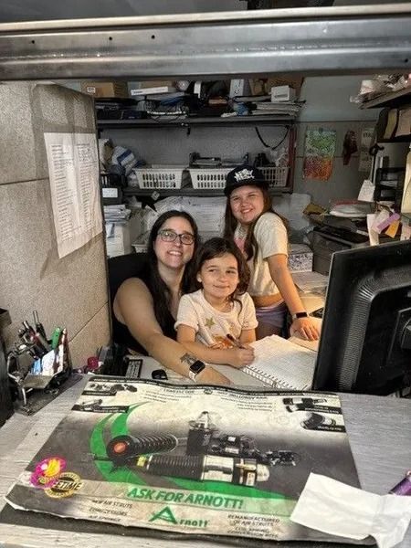 Woman and two young girls smiling behind a cluttered desk with a colorful vintage ad. Setting appears to be an auto shop office.