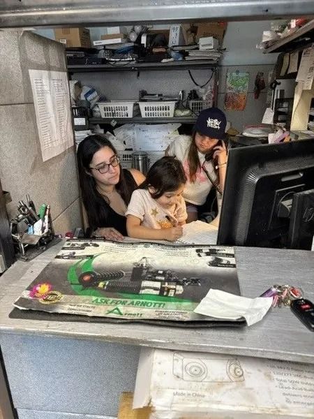 Three people at a counter in a cluttered auto shop; a woman on the phone, a young child drawing, and a woman watching.