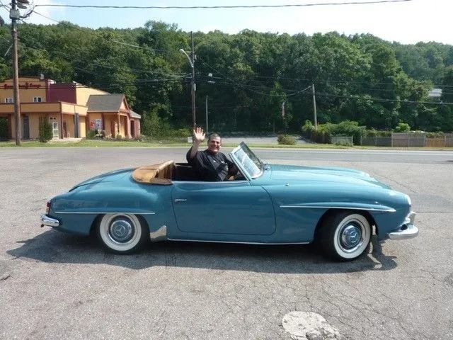 Man waving from a light blue vintage convertible with white-walled tires, parked in front of a building and trees.