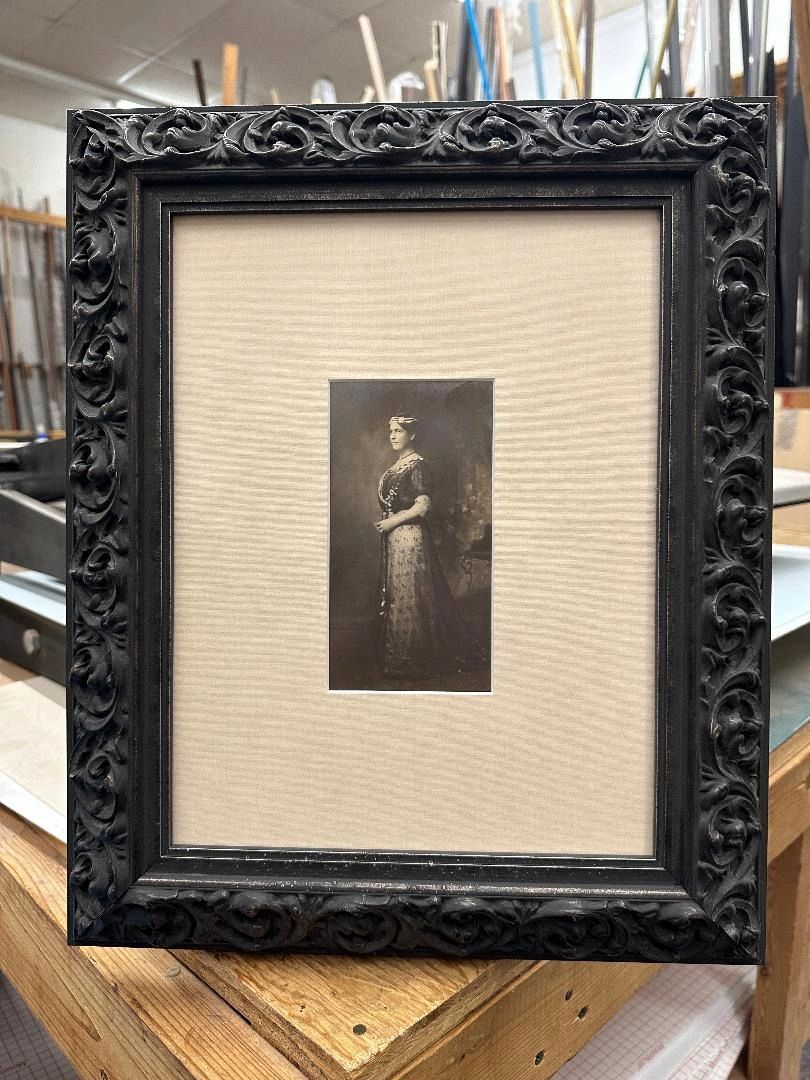 Framed portrait of a woman in black dress, with ornate black frame, set on a beige mat.
