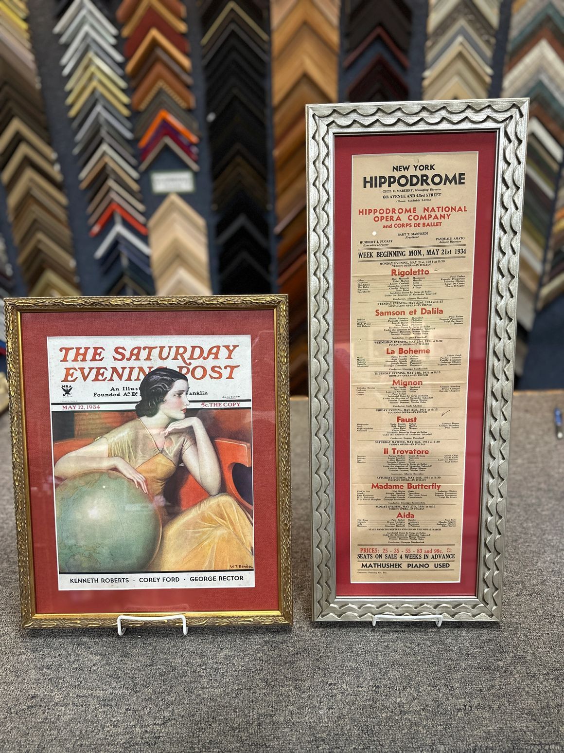Two framed vintage artworks: a magazine cover and a handwritten document, on a display table against a backdrop of frame samples.