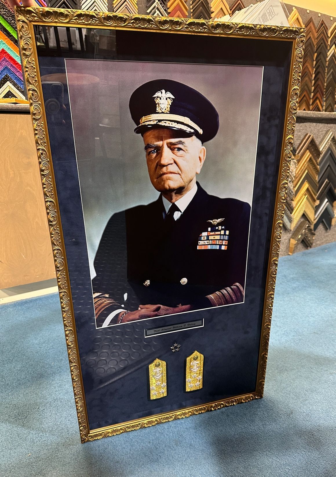 Framed portrait of a naval officer in uniform; gold insignia below the portrait, set against a blue background.