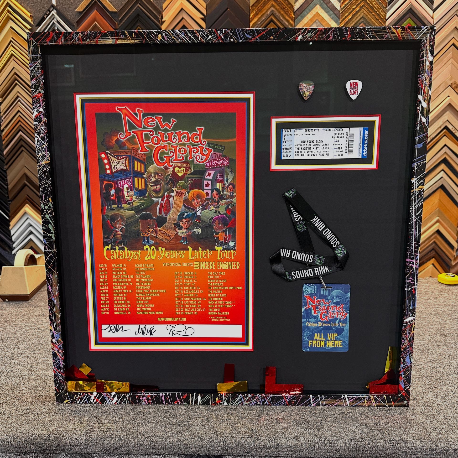 Framed concert memorabilia: poster, ticket, backstage pass, guitar picks, with a colorful border, on a black mat.