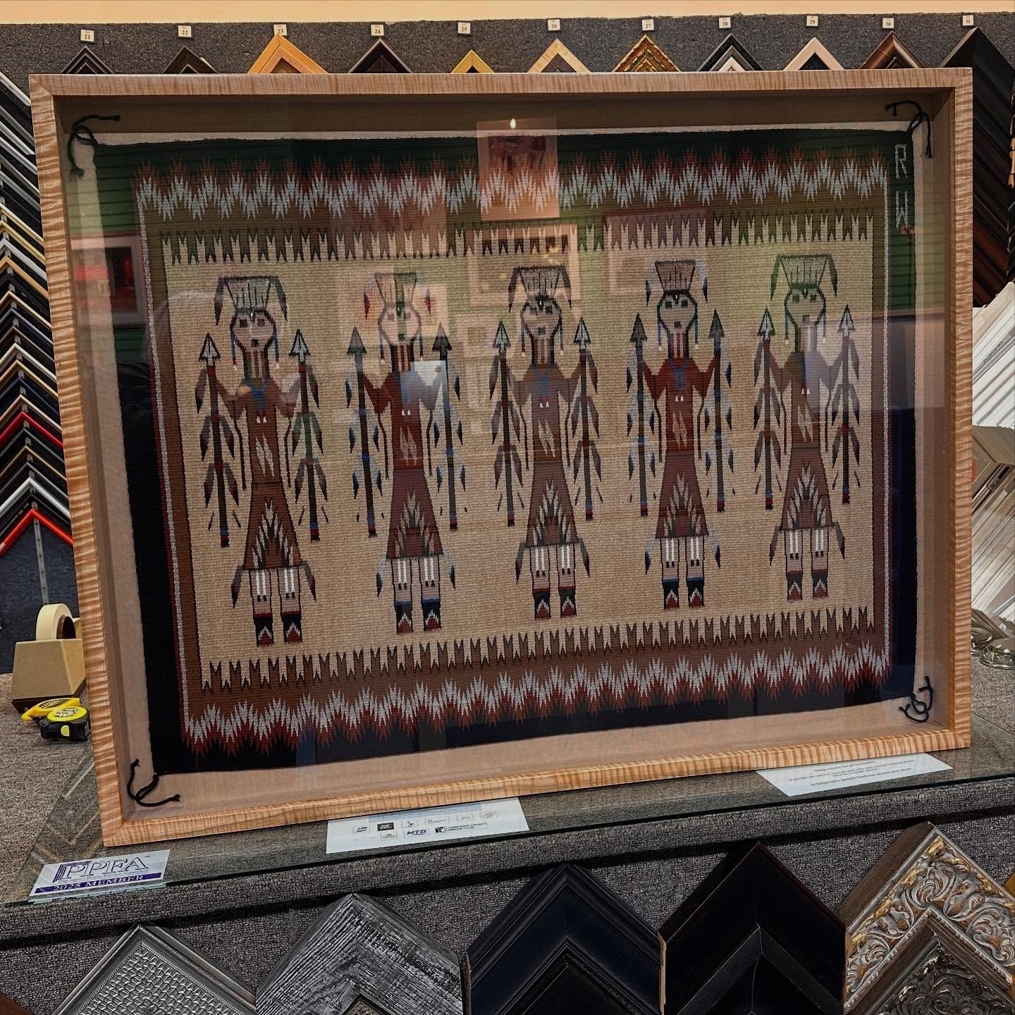 Framed textile art depicting five figures with patterned clothing and raised arms, on display in a shop.