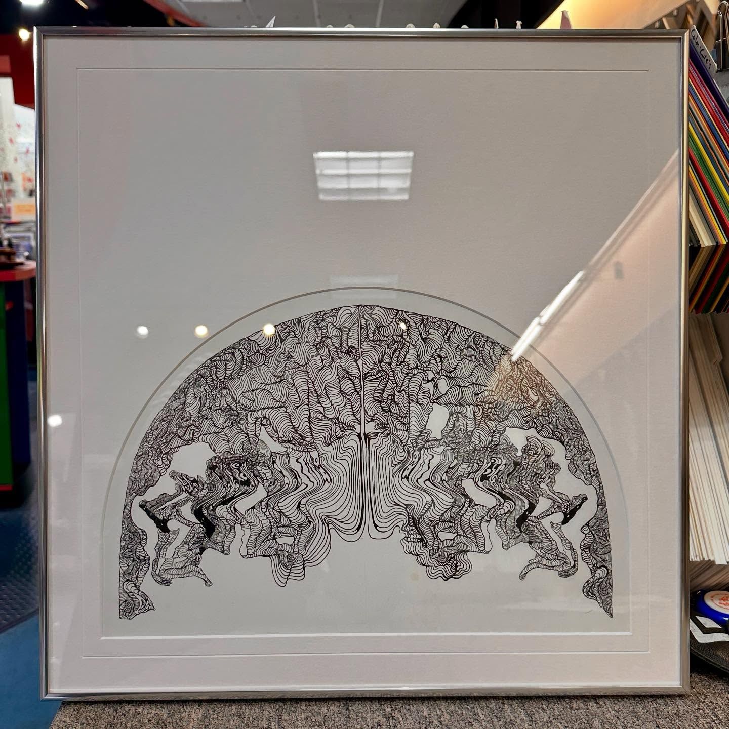 Framed black and white artwork of a tree and figures, in a gallery setting, with a silver frame.