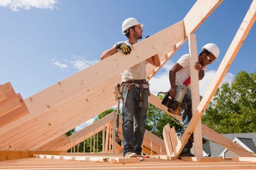 Two construction workers lifting roof trusses on a wooden house frame under a blue sky