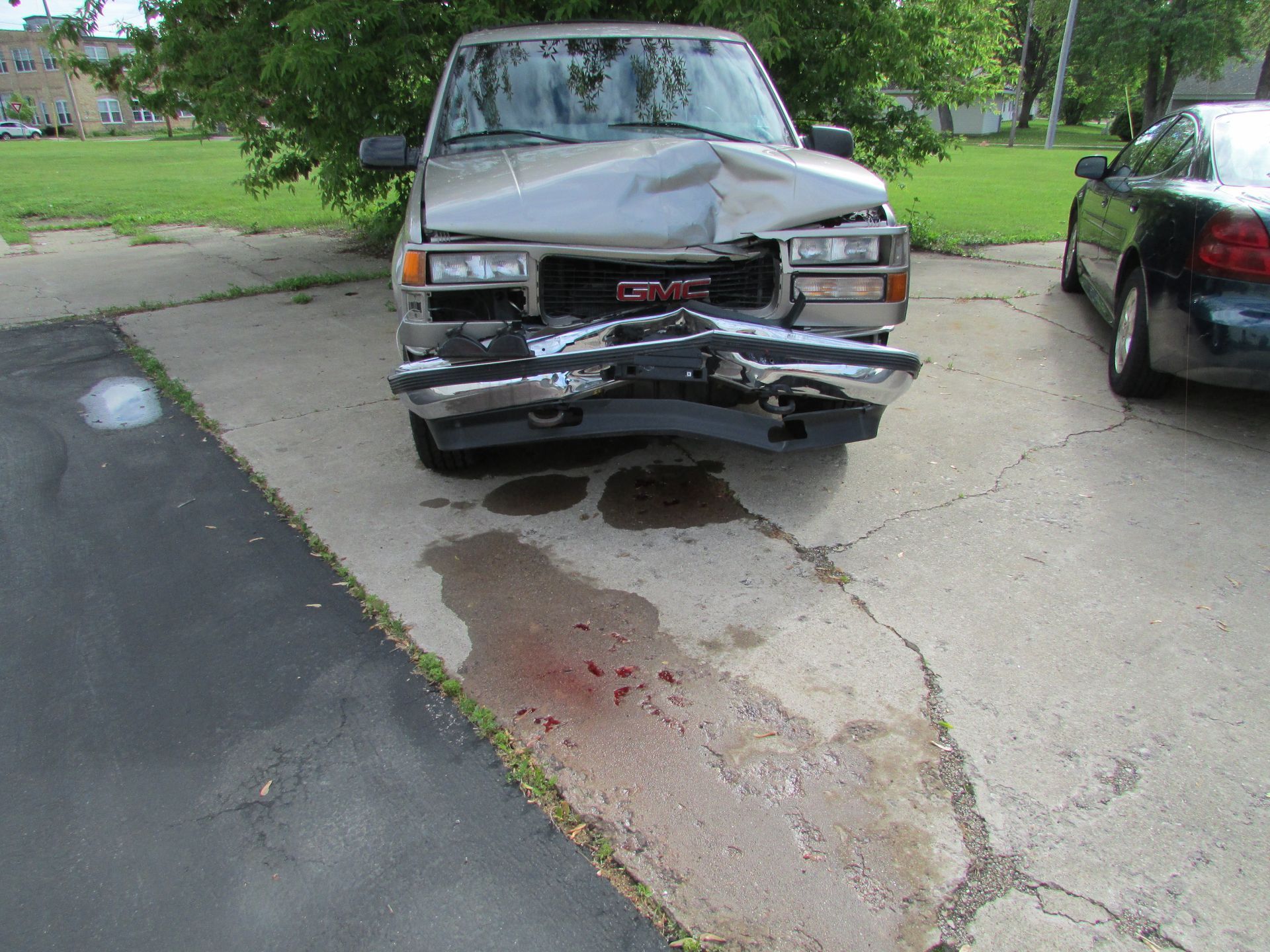 Damaged gray GMC truck front end after a collision, resting on a concrete driveway with a red fluid stain.