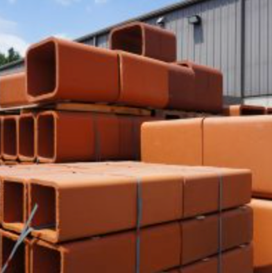 Stacks of orange rectangular metal pipes outdoors beside a warehouse