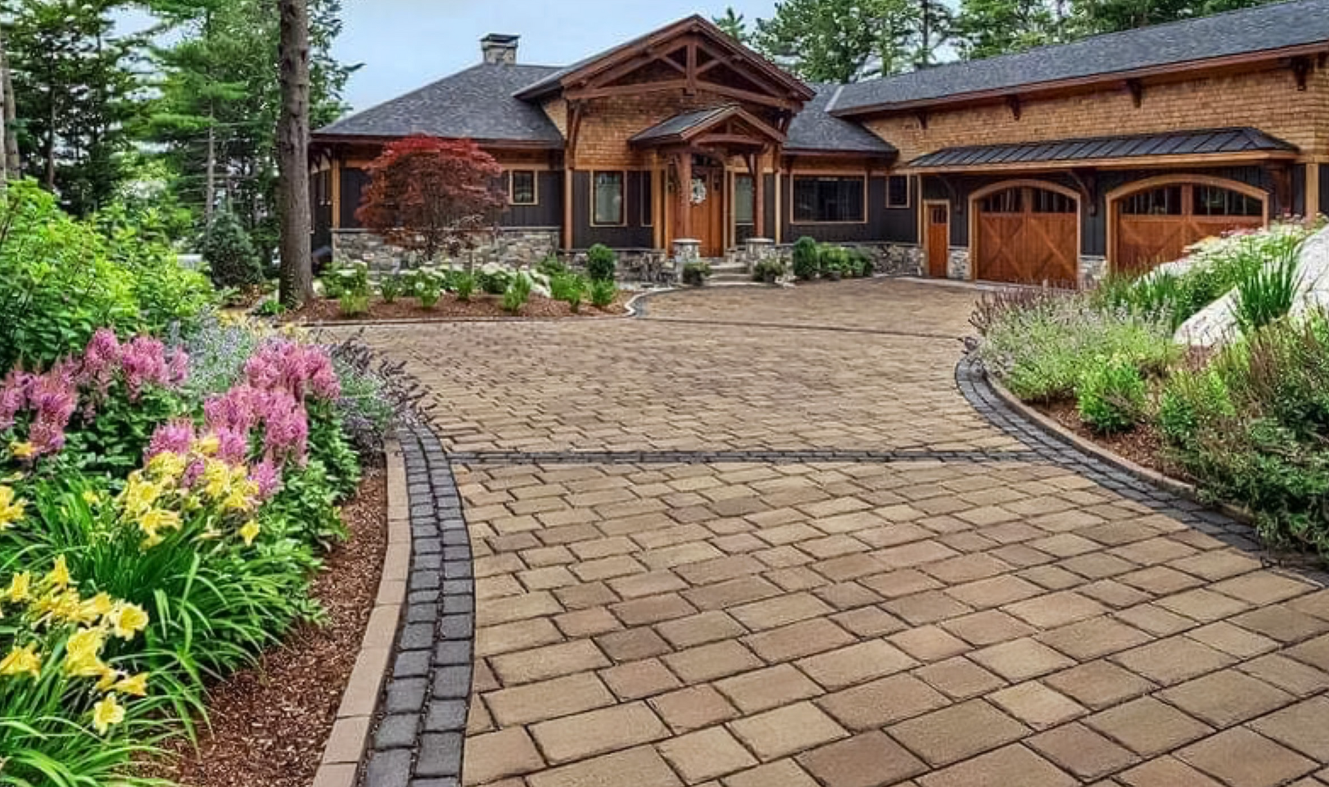 Stone driveway leading to a rustic brick house with a landscaped flower garden and trees.