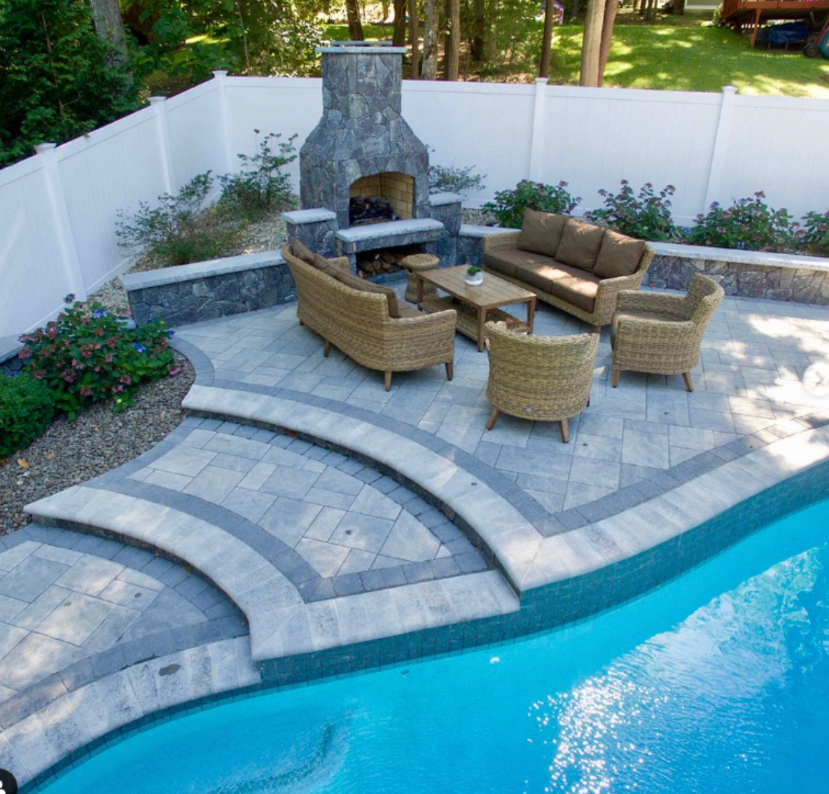 Backyard patio with stone fireplace, wicker seating, and curved pool steps beside a blue swimming pool.