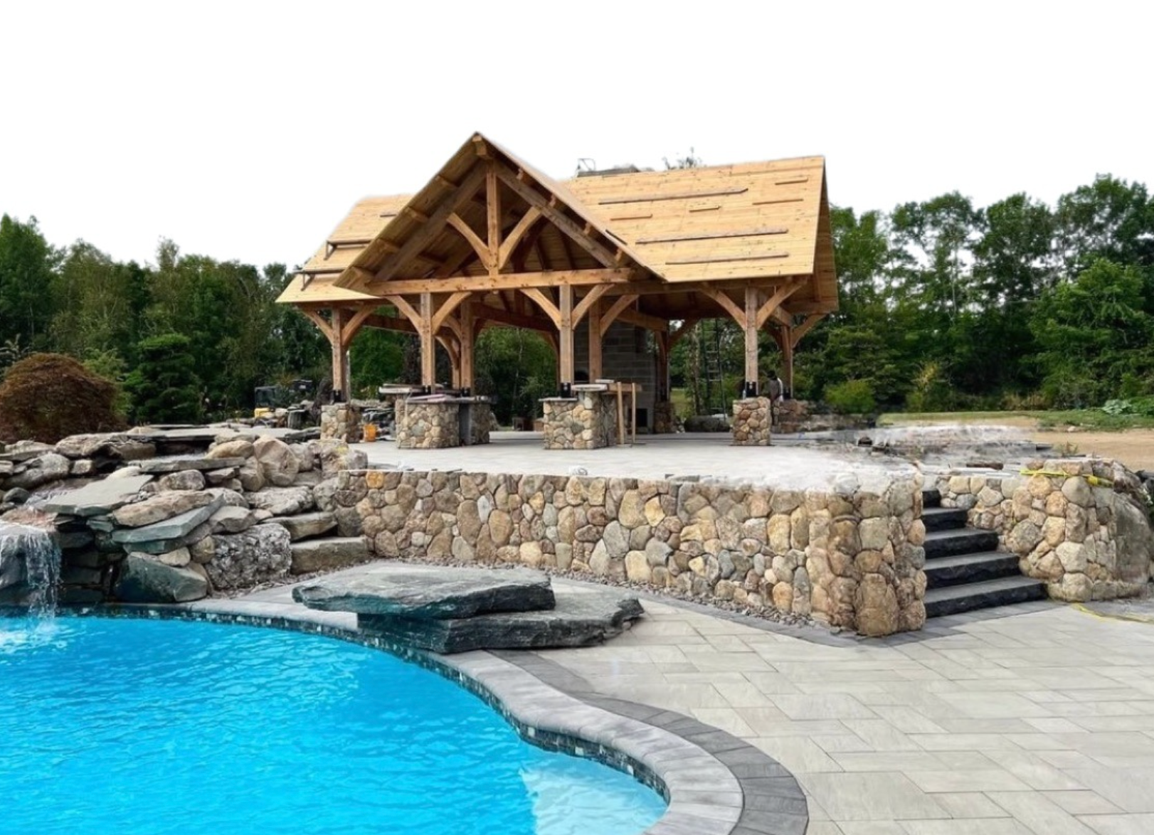 Stone pavilion beside a blue pool, with steps and a rocky landscaped patio in a wooded outdoor setting.