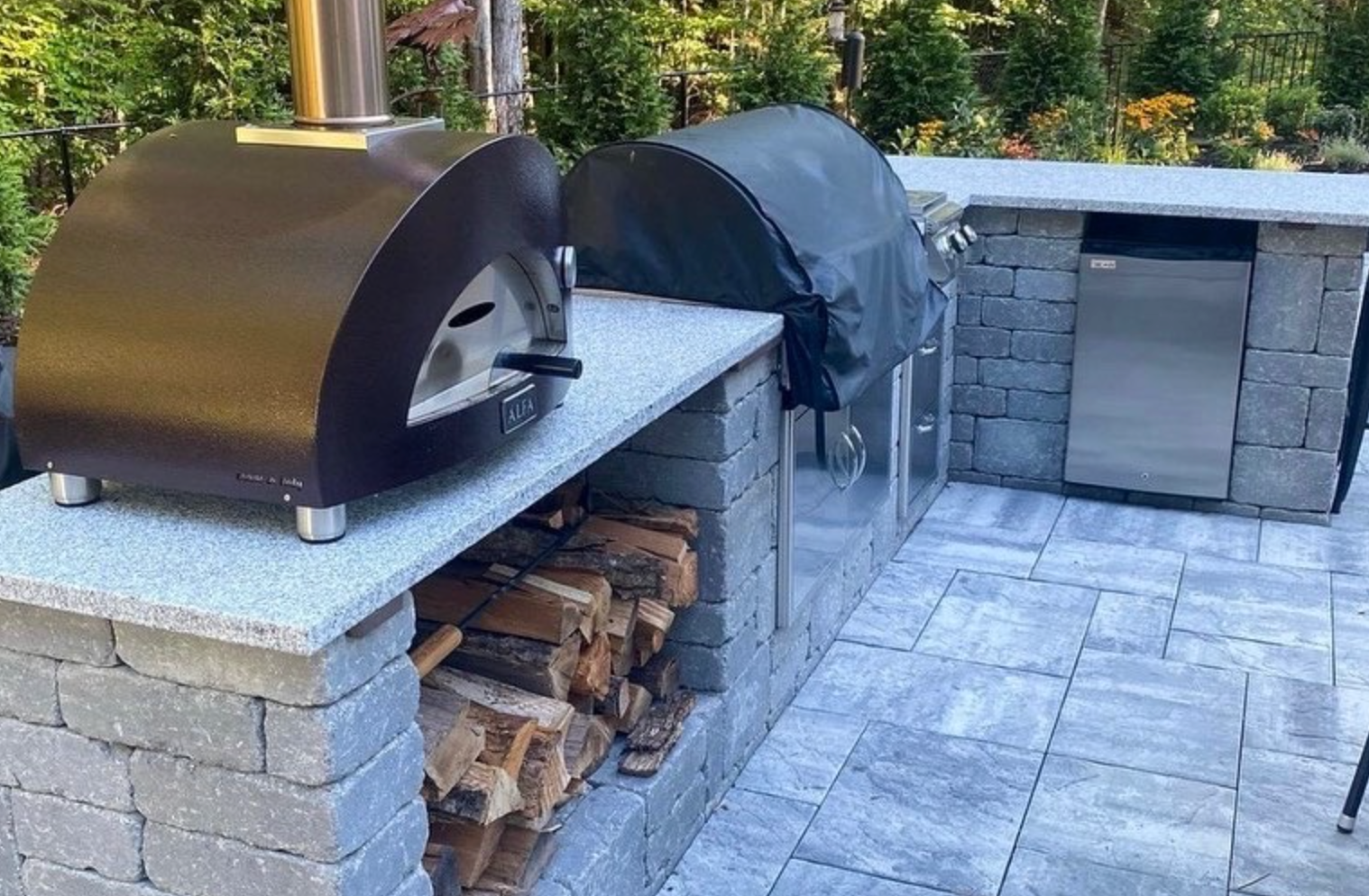 Outdoor stone patio with built-in black and gray pizza ovens and stacked firewood underneath.