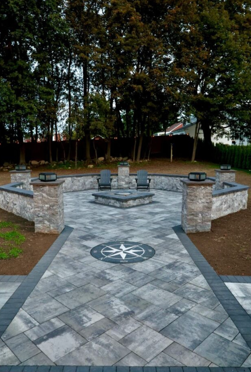 Stone memorial courtyard with benches, a compass rose, and trees in the background.