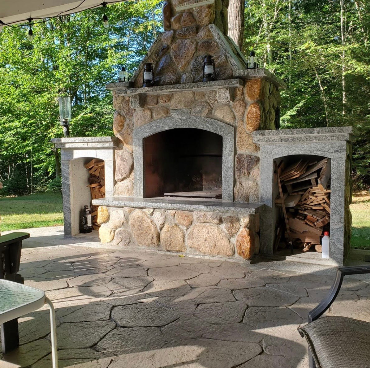 Stone outdoor fireplace with arched firebox, flanked by wood storage niches in a wooded patio.