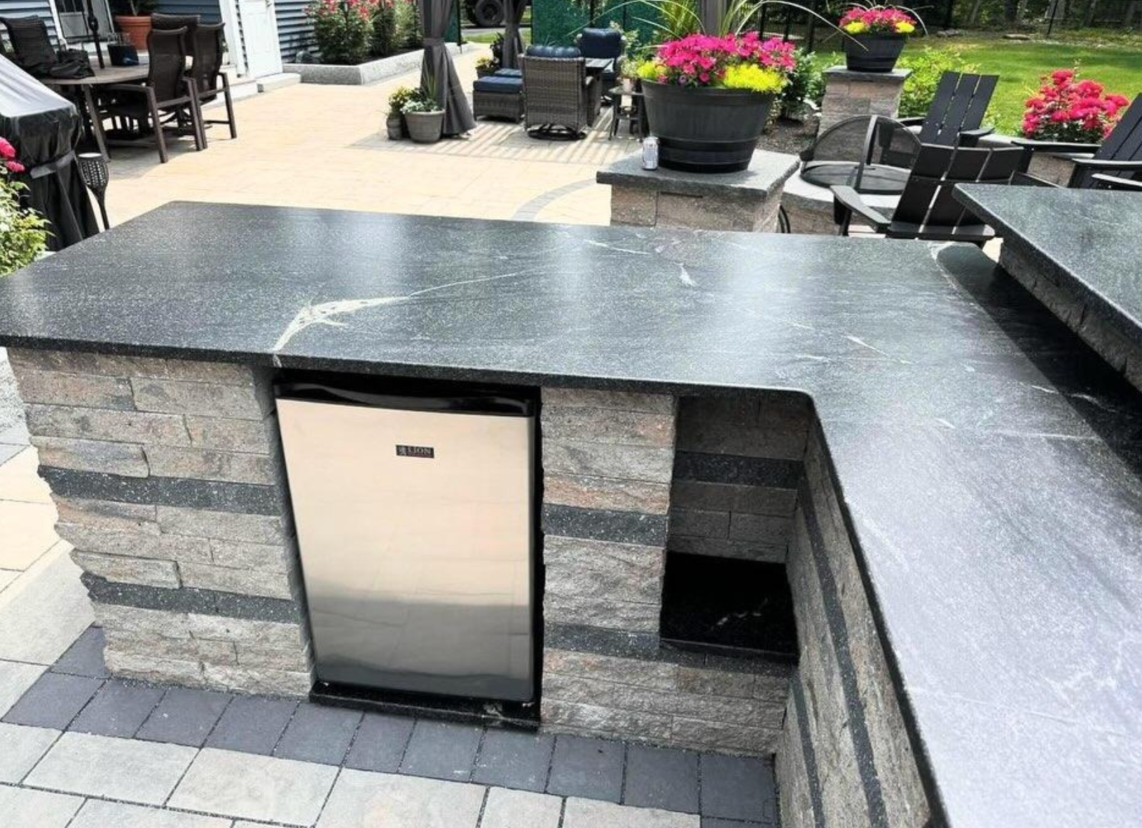 Outdoor stone patio with black countertop and built-in stainless cabinet, surrounded by benches and flowers.