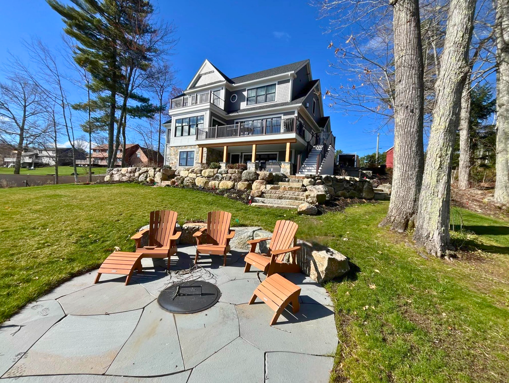 Backyard fire pit seating area with wooden chairs in front of a large gray house on a sunny day.